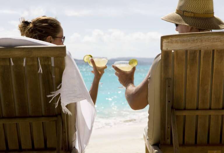 people sit in two wooden beach chairs facing the Caribbean ocean holding drinks and making a toast